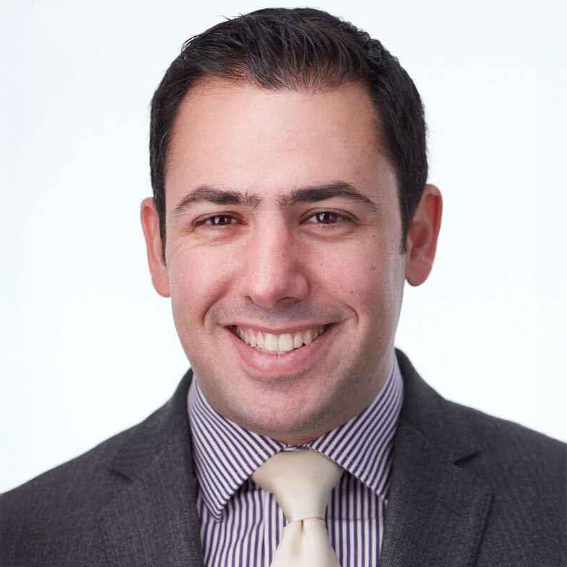 Figure. A man in a grey suit, light tie, and striped shirt smiles at the camera against a white background.